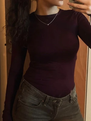 Mörklila långärmad body - Snygg mörklila långärmad topp med tight passform. Toppen har en enkel, stilren design utan synliga detaljer och är perfekt att matcha med jeans eller kjol. Materialet ser mjukt och stretchigt ut för en bekväm känsla. Lila/burgundy