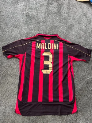 AC Milan fotbollströja Maldini #3 - Säljer en klassisk AC Milan matchtröja från Adidas med Maldini och nummer 3 på ryggen. Tröjan är röd och svart med vertikala ränder, klubbmärke och Champions League-märke på ärmen. Tillverkad i lätt och ventilerande material, perfekt för fotboll eller samling.