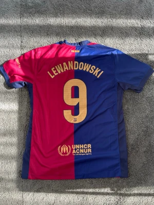 FC Barcelona matchtröja Lewandowski - Säljer en officiell FC Barcelona matchtröja från Nike med Lewandowski och nummer 9 på ryggen. Tröjan är röd och blå, har klubbmärke och sponsorer på framsidan samt UNHCR-logga på baksidan. Perfekt för fotbollsfans som vill visa sitt stöd för laget.