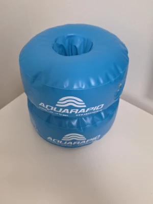 Blåa simringar från Aquarapid - Säljer ett par blåa simringar från Aquarapid, perfekta för simträning eller lek i poolen. Tillverkade i slitstark plast med tryckt logga och hål i mitten för enkel användning. Passar för vikt upp till 30 kg.