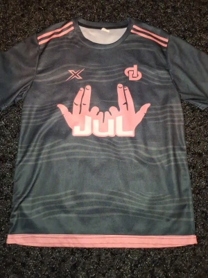 Svart sporttröja med rosa detaljer - Cool svart sporttröja med rosa tryck och detaljer. Framsidan har ett stort motiv med händer och texten 'JUL' samt två loggor på bröstet. Tröjan har rund hals och korta ärmar, perfekt för träning eller sport.