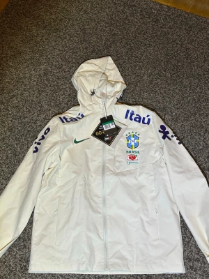 Vit Brasil Nike vindjacka med huva - Snygg vit vindjacka från Nike med Brasilien-landslagets logga och sponsorer tryckta på bröst och ärmar. Jackan har huva med dragsko, hel dragkedja framtill och är tillverkad i slitstarkt GORE-TEX-material. Perfekt för dig som gillar sportig stil.