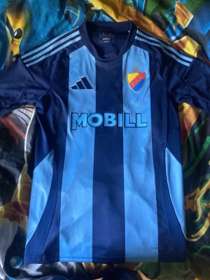 Djurgården matchtröja blå randig - Säljer en blå randig Djurgården matchtröja från Adidas med tryck 'MOBILL' på bröstet och klubbmärke. Nummer 21 och namnet 'Bergvall' på ryggen. Tröjan har korta ärmar och klassiska Adidas-ränder på axlarna. Perfekt för fotbollsfans! Och är även signerad av Lucas bergvall både på ryggen och magen  och är i nytt skick inte använd !