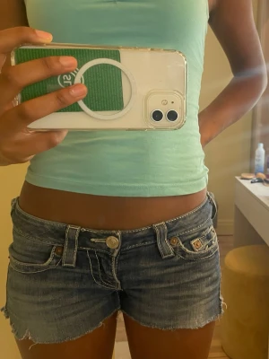 True religion jeans shorts - Säljer ett par snygga lågmidjade jeansshorts i blå denim med slitna kanter och klassiska fickor. Perfekta för varma dagar och en avslappnad stil. Shortsen har knapp- och dragkedjestängning samt detaljer i guldtonad metall.