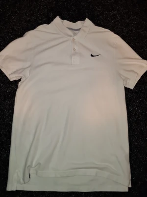 Vit pikétröja från Nike - Klassisk vit pikétröja från Nike med korta ärmar och liten svart logga på bröstet. Tröjan har krage och knappar framtill, perfekt för en clean och sportig look.