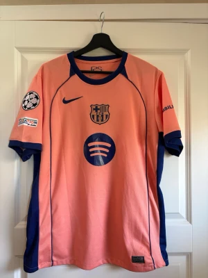 Fotbollströja Barcelona  - Säljer en rosa/orange FC Barcelona matchtröja från Nike med blå detaljer och tryck. Tröjan har klubbmärke, Nike-logga och Spotify som sponsor framtill samt Champions League-märke på ärmen. Perfekt för fotbollsfans!