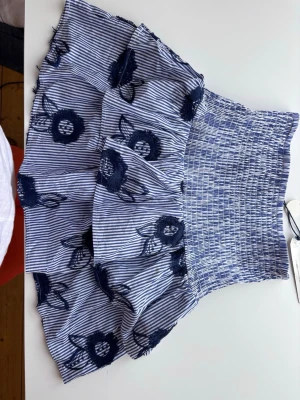 Blåvit kort kjol med broderade blommor storlek S - Säljer en blåvit kort kjol med smala ränder och mörkblå broderade blommor. Kjolen har volanger och bred smock i midjan. Perfekt skick! Oanvänd! Storlek S, från Lindex