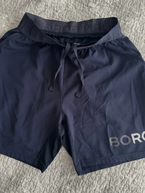 Mörkblå träningsshorts med snörning158/164 cm - Snygga mörkblå träningsshorts med snörning i midjan och elastisk linning. Shortsen har en diskret logga på benet och är tillverkade i ett lätt och snabbtorkande syntetmaterial. Perfekta för sport eller gympass.