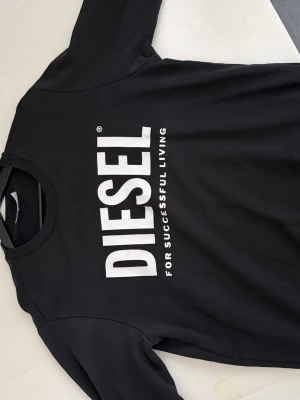 Svart tröja från Diesel - Snygg svart tröja från Diesel med stort vitt tryck på bröstet där det står 'DIESEL FOR SUCCESSFUL LIVING'. Klassisk rund halsringning och rak passform. Perfekt för dig som gillar streetwear och vill ha en enkel men stilren look.