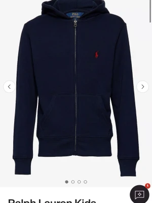 Mörkblå hoodie från Polo Ralph Lauren - Mörkblå hoodie med dragkedja från Polo Ralph Lauren. Klassisk design med huva, fickor framtill och röd broderad logga på bröstet. Tillverkad i mjukt material som känns skönt mot huden. Perfekt för en avslappnad och stilren look.