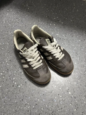 Adidas samba - Säljer ett par klassiska Adidas sneakers i brunt läder och mocka med vita detaljer och snören. Skorna har den ikoniska tre ränder-designen på sidorna och en gummisula. 