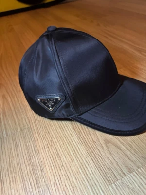 Svart keps från Prada i satin - Snygg svart keps från Prada i glansigt satintyg med klassisk böjd skärm. Kepsen har Pradas ikoniska metallogo på sidan och stilrena sömmar. Perfekt accessoar för en trendig och sportig look.