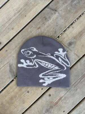 Grå mössa med grodmotiv Beanie  - Säljer en grå mössa med ett stort vitt grodmotiv på framsidan. Mössan är stickad och har en klassisk, rundad form som sitter snyggt på huvudet. Perfekt för dig som vill ha en unik accessoar med coolt djurmotiv.