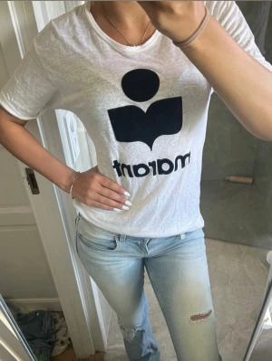 Vit t-shirt från Marant med tryck - Säljer en vit t-shirt från Marant med stort svart logotryck framtill. T-shirten har rund halsringning och korta ärmar. Materialet känns lätt och luftigt, perfekt för en avslappnad stil.