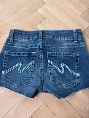 Blå jeansshorts från Nelly - Säljer ett par blå jeansshorts från Nelly med råa kanter och slitna detaljer. Shortsen har fem fickor, bälteshällor och coola broderade mönster på bakfickorna. Perfekta för sommaren och ger en avslappnad vibe.