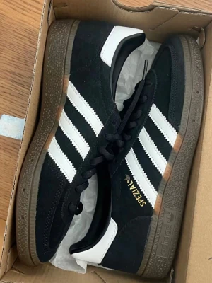 Adidas Spezial svarta sneakers - Säljer ett par Adidas Spezial sneakers i svart mocka med vita detaljer och klassiska tre ränder på sidan. Skorna har brun sula och guldfärgad Spezial-text på sidan. Perfekta för dig som gillar retrostil och streetwear.