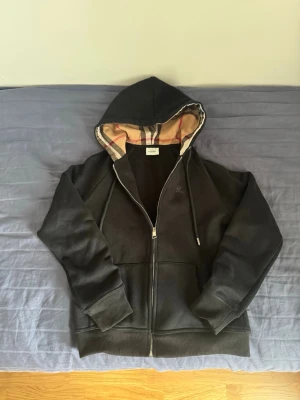 Svart Burberry zip hoodie med dragkedja och huva - Säljer en svart hoodie med dragkedja och klassisk huva. Insidan av huvan har ett beige, rött och svart rutigt mönster. Hoodien har två fickor framtill och ribbade muddar vid ärmar och nederkant. Perfekt för en avslappnad och stilren look. Tröjans storlek är S men den sitter som en SX!