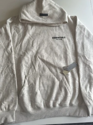 Vit Essentiallss - Snygg beige hoodie från Essentials Fear of God med svart logga på bröstet och stor text på ryggen. Hoodien har en klassisk huva, känguruficka och ribbade muddar. Säljer för jag fick den för stor och inte min stil och kvalité 
