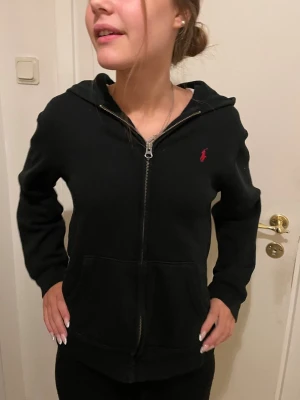 Svart hoodie från Polo Ralph Lauren - Säljer en svart hoodie från Polo Ralph Lauren med dragkedja framtill och klassisk röd logga broderad på bröstet. Hoodien har huva, fickor framtill och är tillverkad i mjukt material. Perfekt för en avslappnad och stilren look.