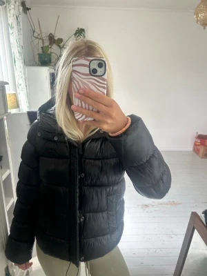 Svart pufferjacka med huva - Säljer en svart pufferjacka med huva och dragkedja framtill. Jackan har två fickor på sidorna och quiltad design som ger en riktigt puffig look. Perfekt för kalla dagar och riktigt skön att bära. Passar dig som gillar en enkel men snygg stil.