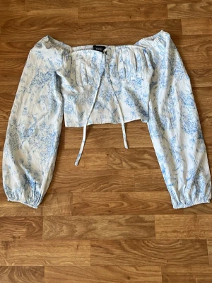 Blommig offshoulder croppad blus - Söt offshoulder blus med blått blommönster på vit botten. Croppad modell med långa puffärmar och smockad rygg. Knytband framtill ger en extra detalj. Perfekt för en trendig och romantisk look.