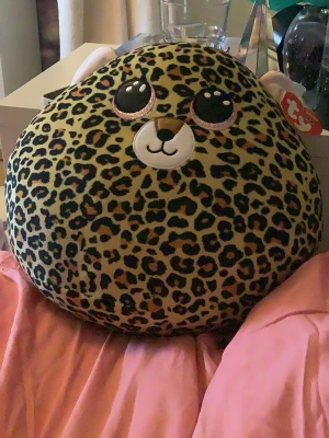 Leopardmönstad mjukisdjur kudde - Supergullig rund mjukisdjur kudde med leopardmönster och stora glittriga ögon. Kudden har små öron, svans och ett broderat ansikte. Perfekt som dekoration eller att krama om. Tillverkad i mjukt plyschmaterial.
