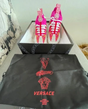 Versace rosa baroque sneakers - Säljer ett par färgstarka Versace sneakers i hot pink och guld med barockmönster. Skorna har vita snören, högt skaft och detaljer i neopren. Ikoniska grekiska mönster på plösen och klassisk Versace-logga. Perfekta för dig som vill sticka ut.