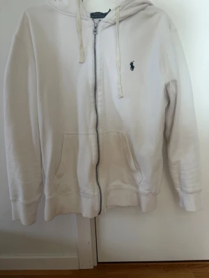 Vit hoodie med dragkedja från Polo Ralph Lauren - Vit hoodie från Polo Ralph Lauren med dragkedja framtill, två fickor och klassisk huva med snören. Broderad logga i marinblått på bröstet. Tillverkad i mjukt bomullsmaterial för en skön och avslappnad look.