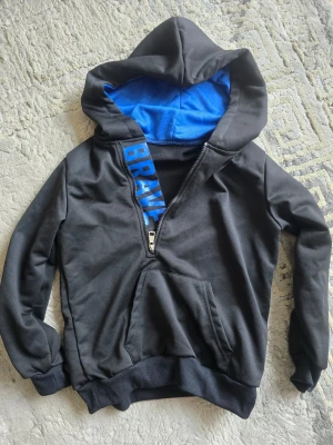 Svart hoodie med blått foder och tryck - Snygg svart hoodie med blått innerfoder i huvan och texten 'BRAVE' i blått längs dragkedjan. Hoodien har känguruficka framtill, ribbade muddar och en halv dragkedja. Perfekt för en avslappnad och cool stil.