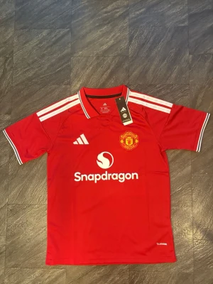 Manchester United matchtröja röd - Snygg röd Manchester United matchtröja från Adidas med vita detaljer och klubbmärke på bröstet. Tröjan har klassiska vita ränder på axlarna, krage och Snapdragon-logga framtill. Perfekt för fotboll och träning.
