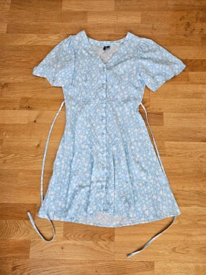 Blommig ljusblå klänning Vero Moda - Söt ljusblå kortklänning från Vero Moda med vitt blommönster. Klänningen har v-ringning, knappar framtill, korta ärmar och knytband i midjan för en feminin siluett. Perfekt för varma dagar och somriga tillfällen.