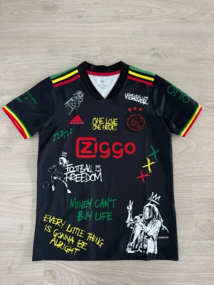 Ajax x Bob Marley Adidas tröja - Säljer en svart Ajax x Bob Marley matchtröja från Adidas med färgglada detaljer i rött, gult och grönt. Tröjan har tryck som 'One Love', 'Football is Freedom' och illustrationer inspirerade av reggae. Materialet är lätt och andas, perfekt för fotboll eller streetwear.