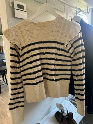 Beige stickad tröja med volang - Söt beige stickad tröja med svarta ränder och volangdetaljer vid axlarna. Tröjan har kabelstickade partier och ribbade muddar vid ärmslut och nederkant. Rund halsringning och mjukt material.