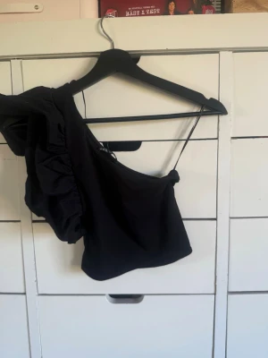 Svart oneshoulder topp från Zara - Säljer en svart oneshoulder topp från Zara med volangdetalj på ena axeln. Toppen har en smal axelrem och är croppad i modellen, perfekt för att lyfta vilken outfit som helst.