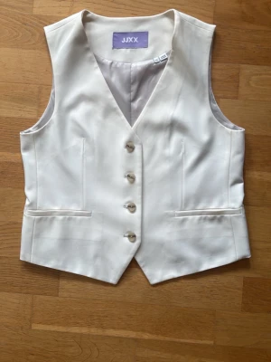 Vit väst från JJXX med knappar - Stilren vit väst från JJXX med fyra knappar framtill och två fickor. Västen har en klassisk v-ringad design och är tillverkad i ett slätt material som ger en elegant look. Perfekt att styla över en skjorta eller t-shirt för en trendig outfit.