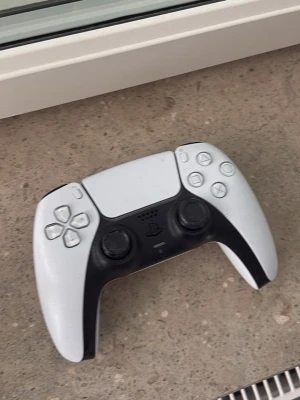 Vit och svart PlayStation-handkontroll - Säljer en vit och svart PlayStation-handkontroll med klassisk design och tydliga knappar. Handkontrollen har två analoga spakar, styrkors och touchpad. Perfekt för gaming på konsol och har en modern look med ergonomisk form.