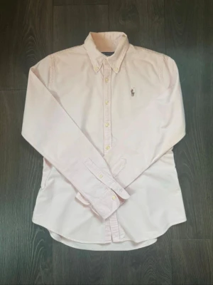  Ralph Lauren skjorta  - Klassisk rosa skjorta från Ralph Lauren med broderad logga på bröstet. Skjortan har knappar framtill, button-down krage och långa ärmar. Perfekt för en clean och stilren look.