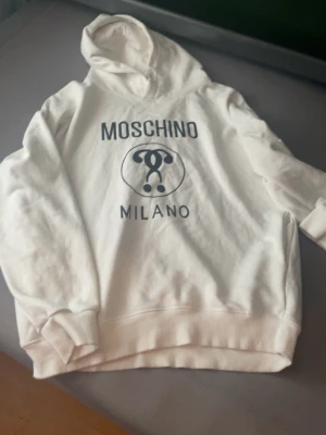 Vit hoodie från Moschino Milano - Snygg vit hoodie från Moschino Milano med stor svart logga och text på bröstet. Klassisk passform med huva och ribbade muddar vid ärmar och nederkant. Perfekt för dig som vill ha en trendig och bekväm look.