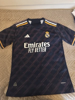 Mörkblå Real Madrid t-shirt - Snygg mörkblå Real Madrid t-shirt med gulddetaljer och vita ränder på axlarna. Adidas-logga och klubbmärke på bröstet samt 'Emirates Fly Better' tryckt framtill. Tillverkad i ett lätt och sportigt material, perfekt för fotboll eller träning. OBS: Tröjan är INTE äkta, därav lågt pris. 