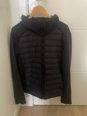 Svart dunjacka med huva - Snygg svart dunjacka med huva och dragkedja framtill. Jackan har quiltade paneler och släta ärmar för en modern look. Perfekt för kalla vinterdagar och enkel att matcha med olika outfits. Skön jackkofta perfekt för hösten/våren! Priset är förhandlingsbart!