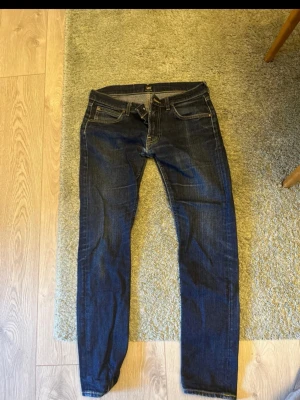 Mörkblå slim fit jeans Lee - Snygga mörkblå slim fit jeans från Lee med klassisk femficksdesign och kontrastsömmar. Jeansen har en rak passform och är tillverkade i denim med lätt stretch för extra komfort. Perfekta för en avslappnad och trendig stil.