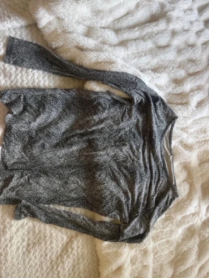 Glittrig långärmad topp - Snygg långärmad topp i mörk silverfärg. Toppen har en figurnära passform och är perfekt för dig som vill sticka ut lite extra. Materialet känns mjukt och stretchigt, vilket gör den bekväm att bära. Passar perfekt till ett par jeans eller kjol.