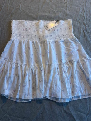 Ny med prislapp, Vit blommog kjol med smock i midjan - Supersöt vit  kortkjol med små blommor och volangdetaljer. Kjolen har en bred smock i midjan som ger en bekväm och snygg passform. Perfekt för varma dagar och enkel att styla med en enkel topp. Helt ny med prislapp kvar.