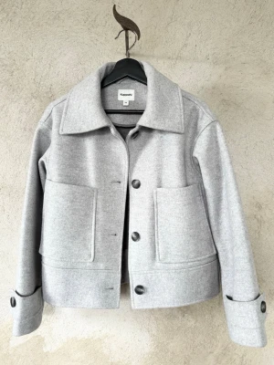 Ljusgrå kort jacka från Kappahl - Stilren ljusgrå kort jacka från Kappahl med stora framfickor, klassisk krage och svarta knappar. Overshirten har raka ärmar med knappdetalj vid mudden och är tillverkad i ett mjukt, filtliknande material som ger en clean och modern look. 