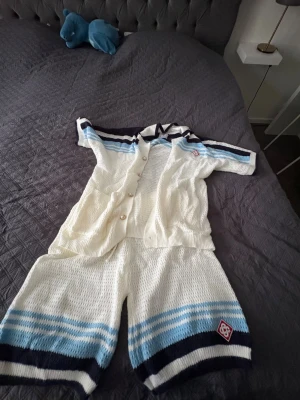 Stickat set i vitt med blå ränder - Säljer ett stickat tvådelat set i vitt med blå och marinblå ränder. Setet består av en kortärmad tröja med knappar och krage samt matchande shorts. Materialet är stickat och luftigt, perfekt för en avslappnad stil.
