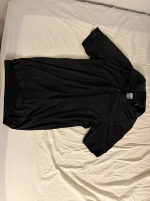 Svart sportpiké från Callaway - Snygg svart sportpiké från Callaway med korta ärmar och knappslå framtill. Tillverkad i ett lätt och snabbtorkande material som passar perfekt för träning eller golf. Diskret logga på ärmen för en clean look.