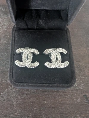 Silverfärgade cc örhängen med stenar - Snygga cc örhängen i silverfärg med den klassiska dubbel-C formen, dekorerade med små gnistrande stenar. Perfekta för dig som vill ha en elegant och trendig accessoar som sticker ut. Kommer i en svart ask.