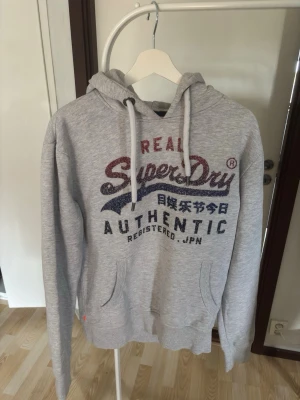 Grå hoodie från Superdry - Säljer en grå hoodie från Superdry med stor logga och japansk text på bröstet. Tröjan har huva med vita snören, känguruficka och mjukt fleecefoder på insidan. Perfekt för dig som gillar streetwear och vill ha något bekvämt och snyggt.