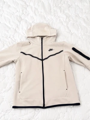 Vit nike tech kofta - Snygg vit vindjacka från Nike med svart dragkedja och detaljer. Jackan har huva, två fickor framtill och en extra ficka med dragkedja på ärmen. Tillverkad i ett slätt material som känns sportigt och modernt. Perfekt för dig som gillar streetwear.