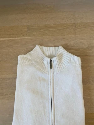 Vit stickad ziptröja från Buddha - Säljer en stilren vit stickad tröja från Buddha med dragkedja framtill och hög ribbad krage. Perfekt för lager på lager och enkel att matcha med olika outfits. Materialet känns mjukt och bekvämt mot huden.